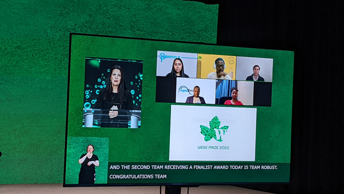 Team reps await Wege Prize competition results, on TV monitor.